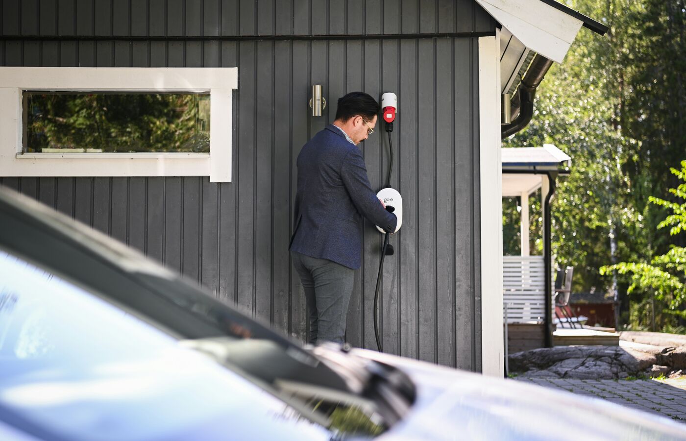 EV driver plugging a charging cable into a wall-mounted home charger in a neighbour's driveway.